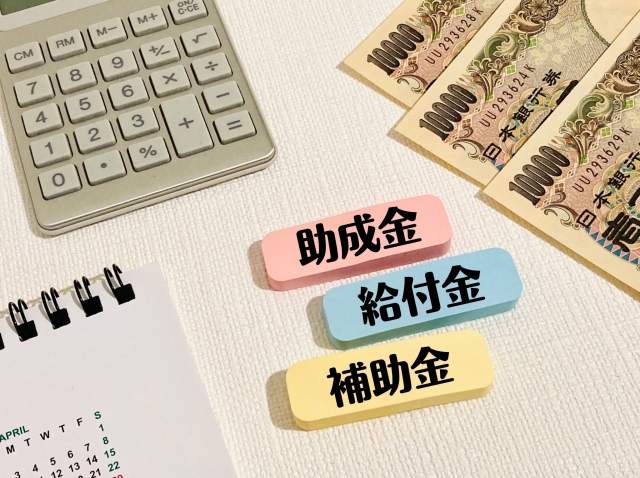 お金と電卓のイメージ写真。助成金や給付金・補助金の名前入り